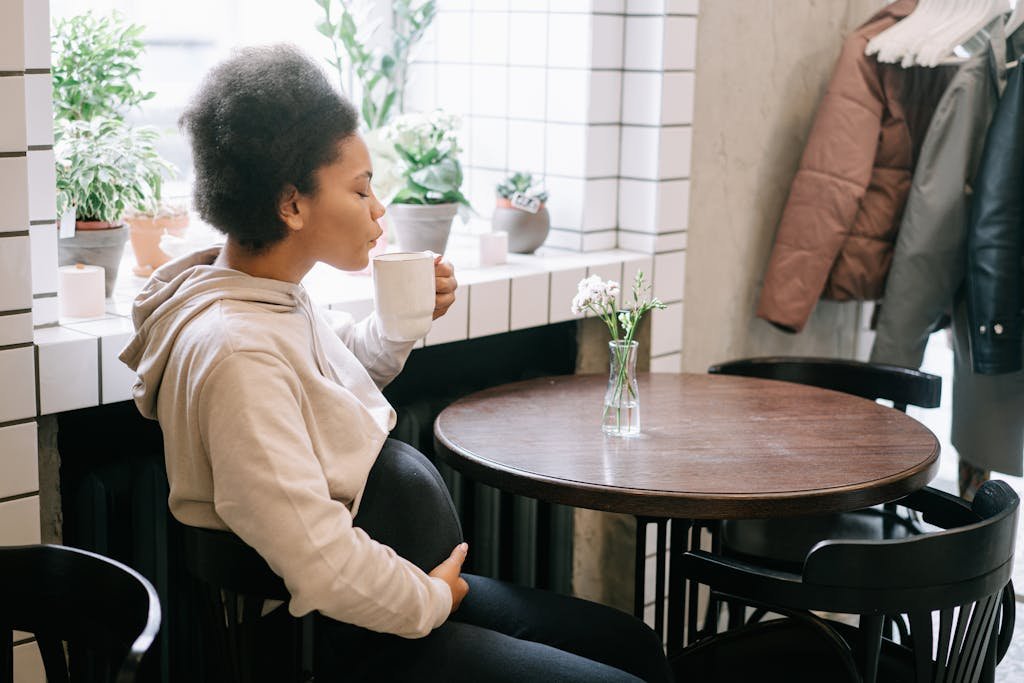 Pregnant woman relaxing with coffee in a cozy café setting, surrounded by plants.